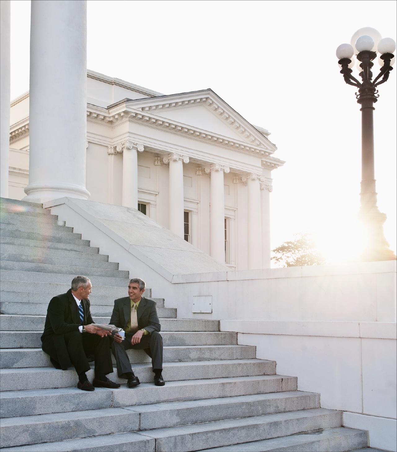Two men in suits sit on the steps outside a neoclassical government building at sunset.